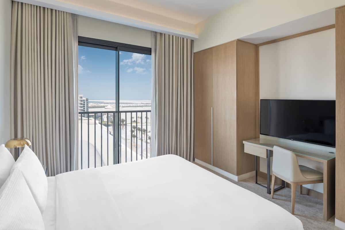 A hotel room with a large bed, a desk with a TV, and a balcony with a view of the city.