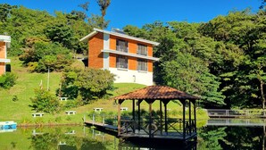 A modern two-story building with a wooden facade, a gazebo by a calm lake, and lush greenery in the background. - Burbi Lake Lodge (Cabeceras)