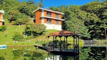 Ein modernes, zweistöckiges Gebäude mit Holzfassade, ein Pavillon an einem ruhigen See und üppiges Grün im Hintergrund.