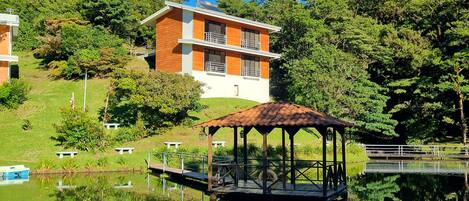 Un edificio moderno su due piani con facciata in legno, un gazebo affacciato su un lago calmo e vegetazione lussureggiante sullo sfondo.