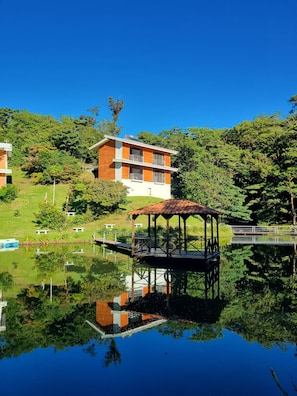 A modern two-story building with a wooden facade, a gazebo by a calm lake, and lush greenery in the background. - Burbi Lake Lodge (Cabeceras)