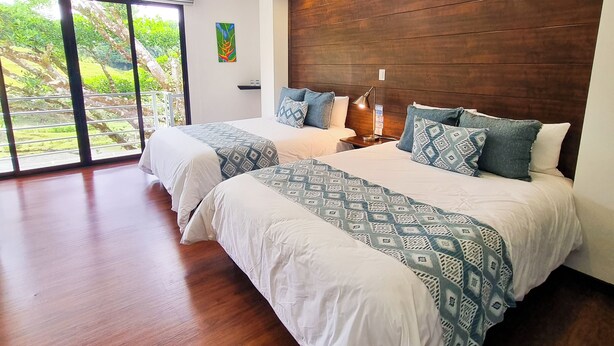 A modern bedroom with a large bed, wooden headboard, and a view of greenery through large windows. - Burbi Lake Lodge (Cabeceras)