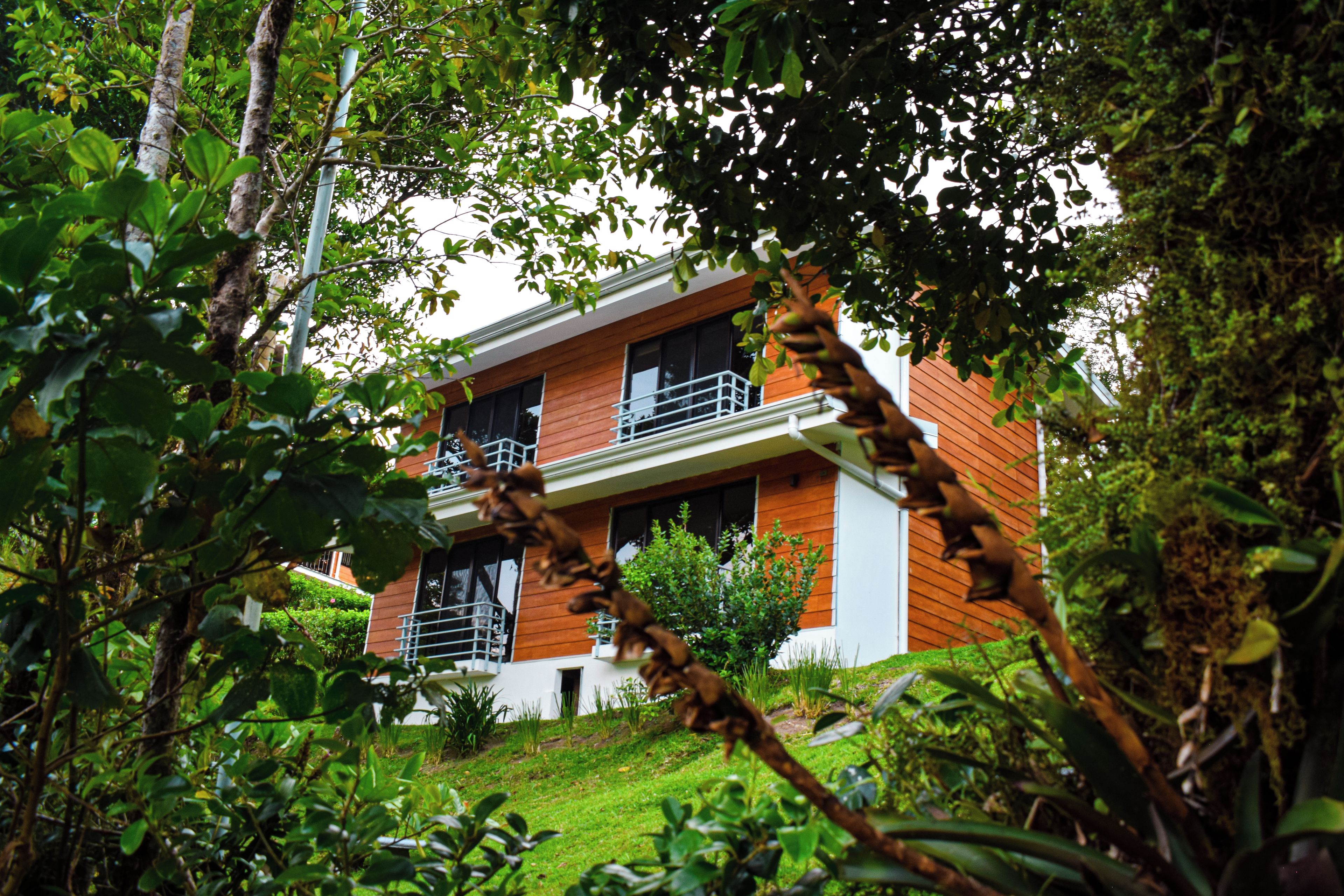 A modern two-story house with a balcony on the second floor, surrounded by lush greenery.