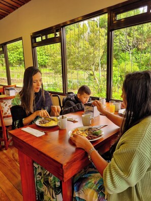 A dining area with a wooden table, chairs, and large windows with a view of greenery. - Burbi Lake Lodge (Cabeceras)
