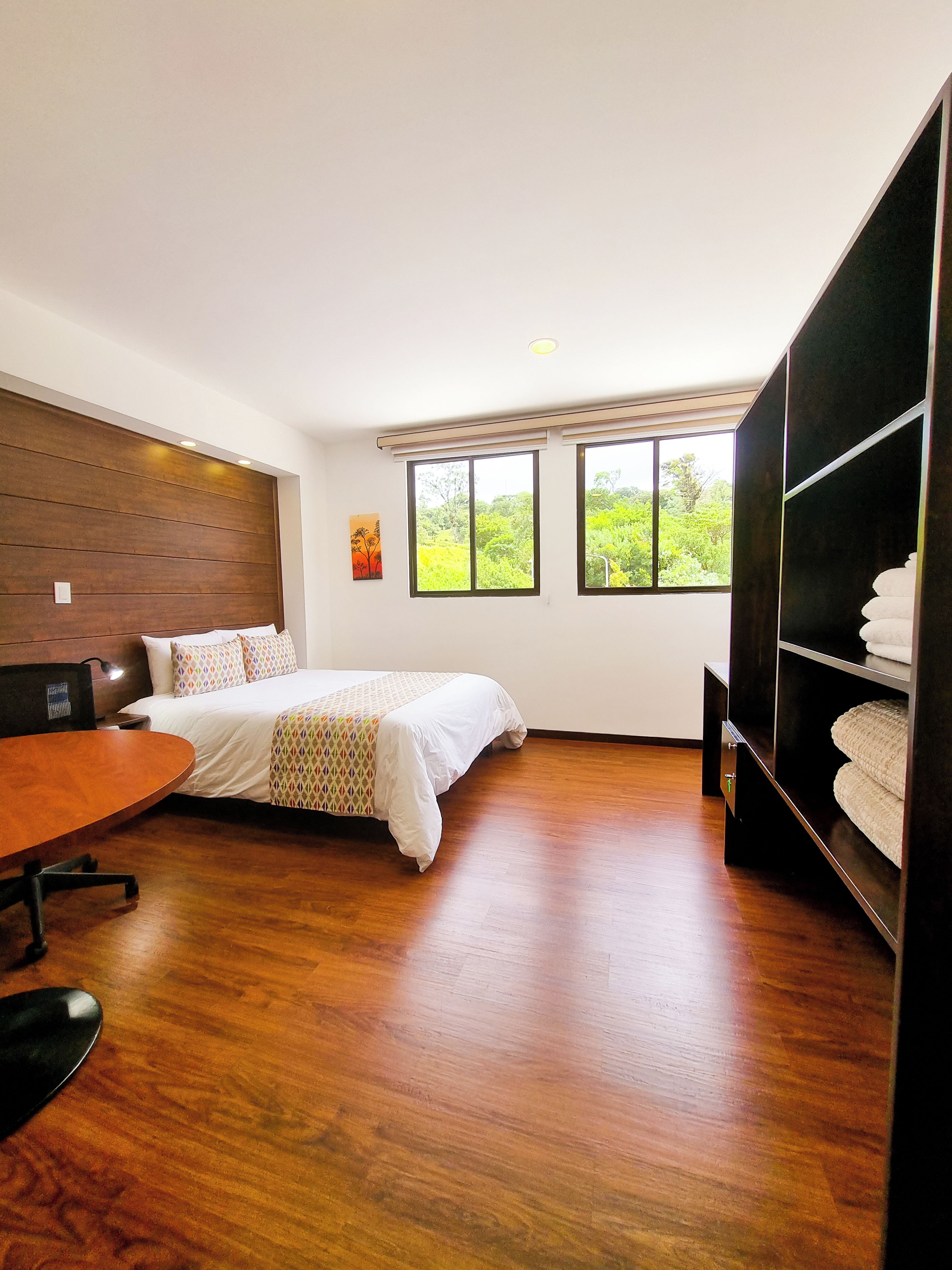 Une chambre d’hôtel équipée d’un lit, d’un bureau, d’une chaise, d’une armoire et de grandes fenêtres donnant sur un paysage verdoyant.