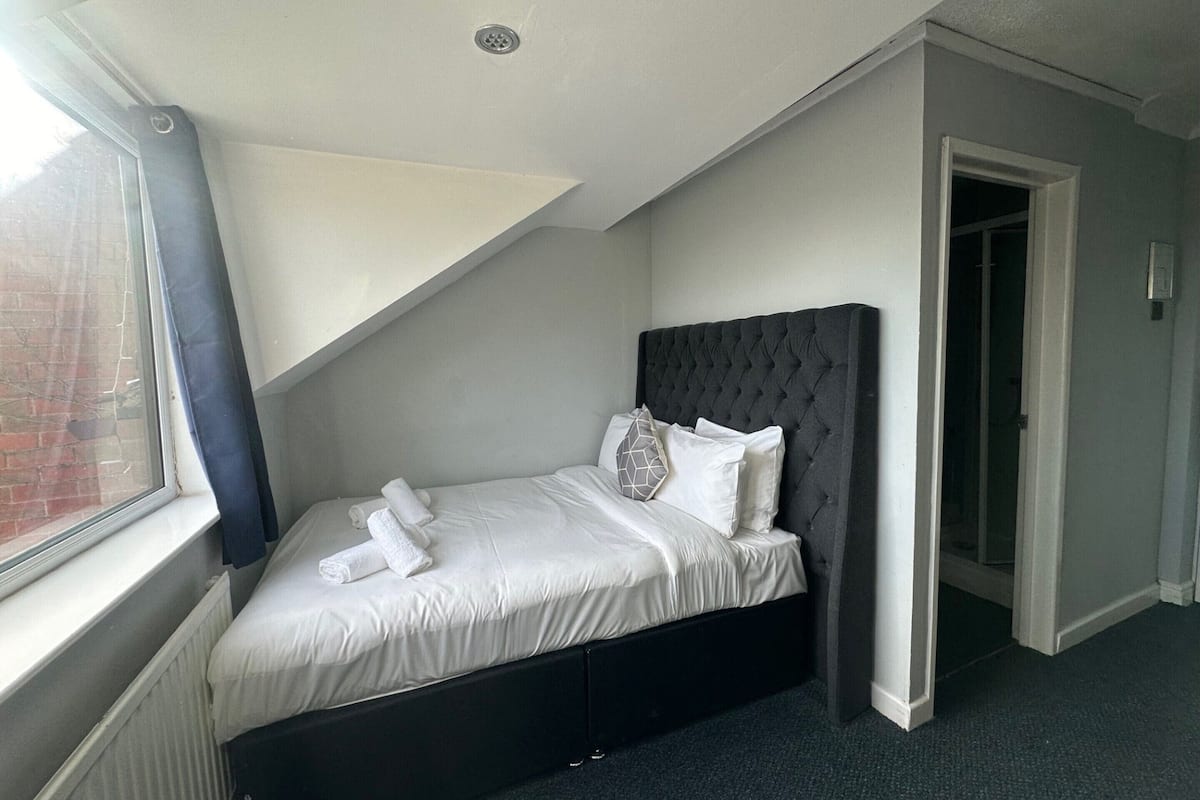 A neatly made bed with white linens and pillows, a dark headboard, and a window with curtains.