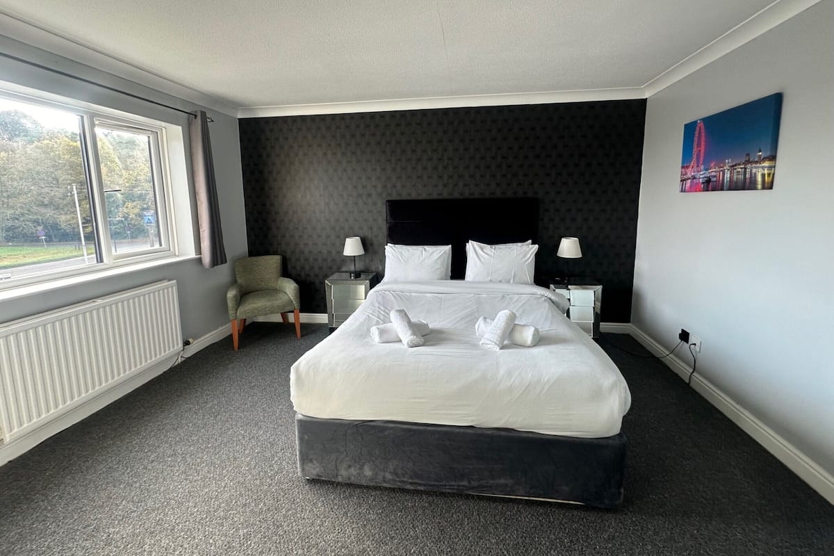 A hotel room with a large bed, two bedside tables with lamps, a chair, a radiator, and a window with a view of trees and a building.