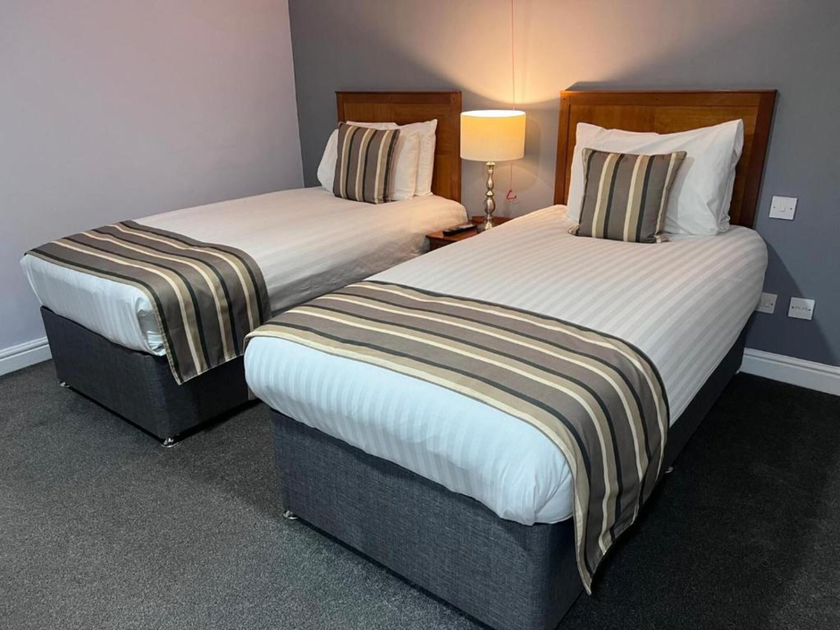 A hotel room with two single beds, each with a wooden headboard, a bedside table, and a lamp.