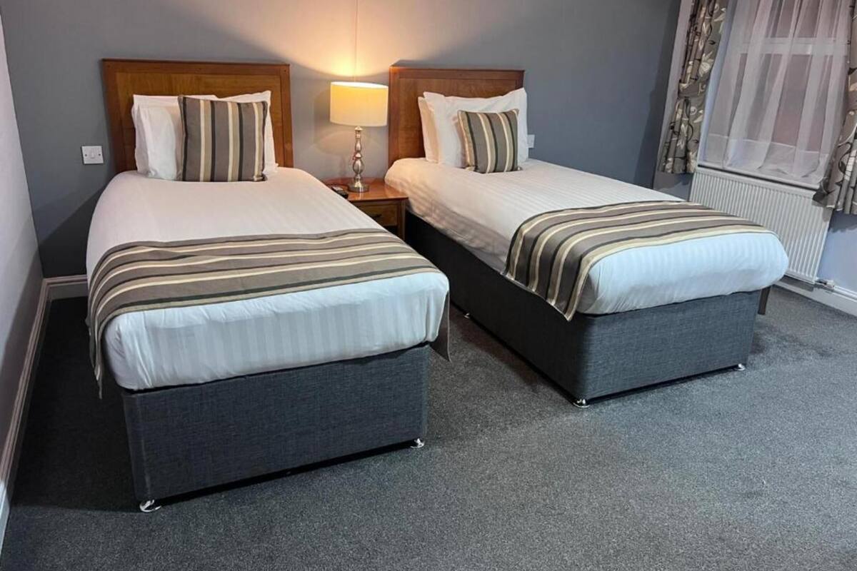 Une chambre d’hôtel avec deux lits simples, chacun doté d’une tête de lit en bois, d’une lampe de chevet et d’une fenêtre avec des rideaux légers.