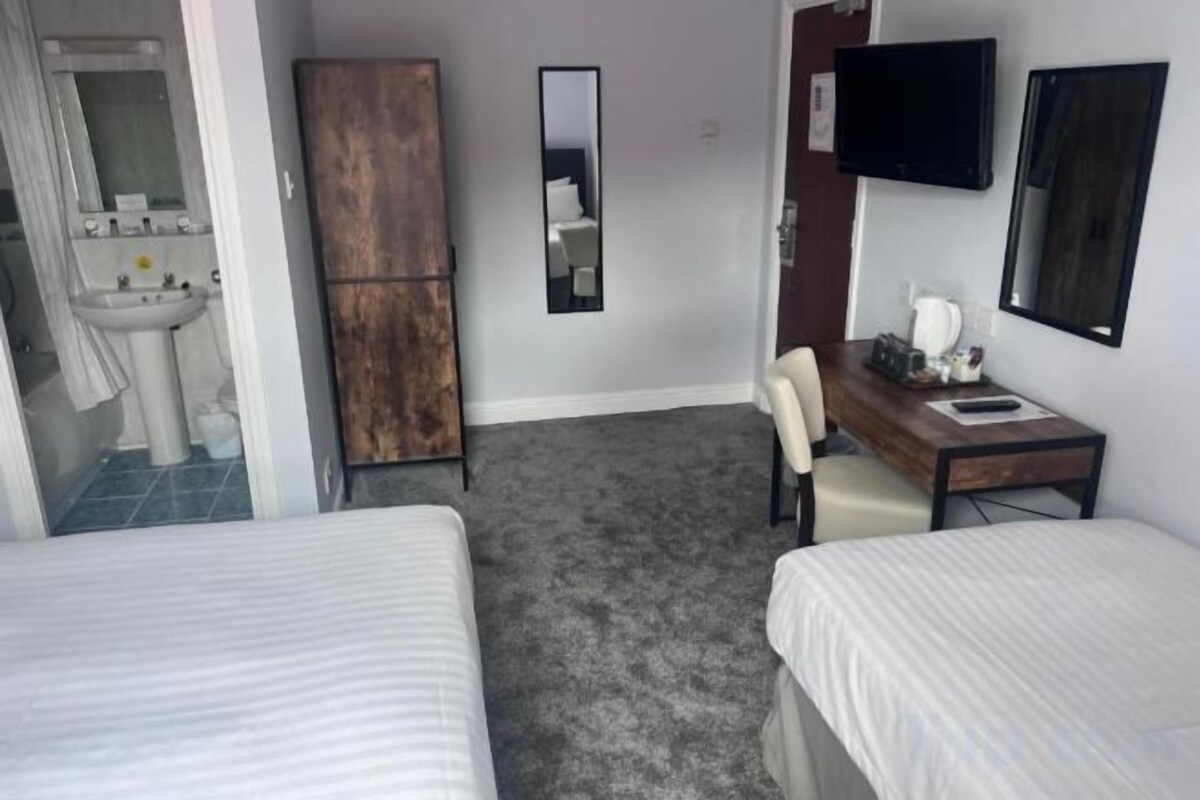 Ett hotellrum med två sängar, ett skrivbord, en stol, en spegel, en tv och ett badrum med handfat och spegel.