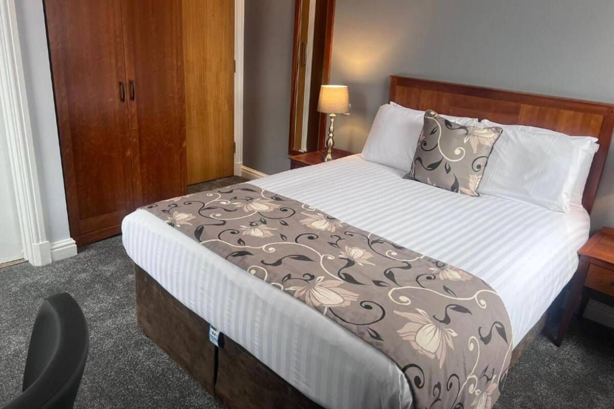 Une chambre d’hôtel avec un grand lit, une armoire en bois, une table de chevet avec une lampe et un sol recouvert de moquette.