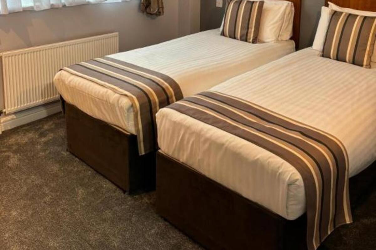 Une chambre d’hôtel avec deux lits, un radiateur, une fenêtre avec des rideaux et un sol recouvert de moquette.