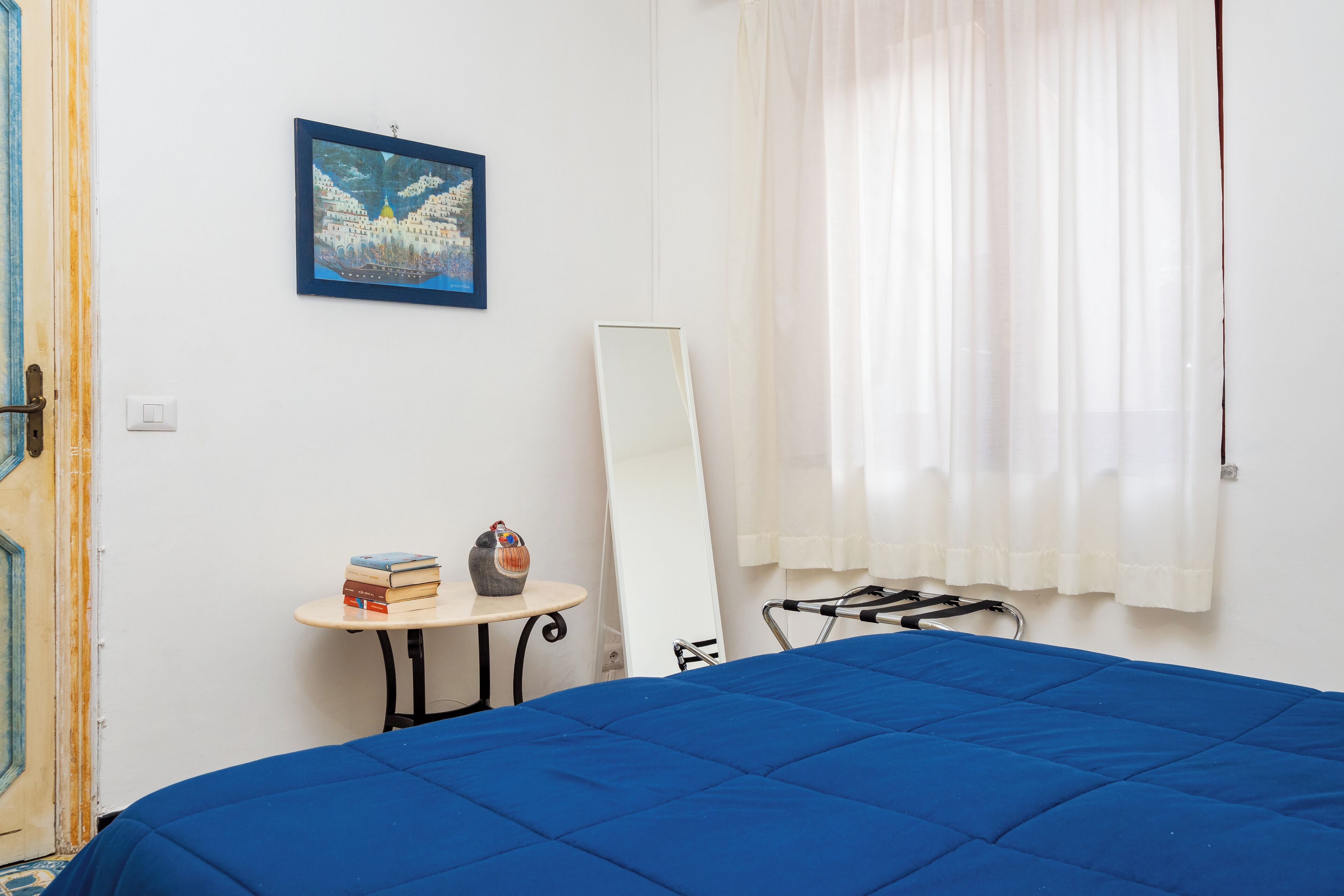 Een slaapkamer met een blauw beddegoed, een klein tafeltje met boeken en een decoratief object, een schilderij aan de muur, een spiegel en een raam met witte gordijnen.