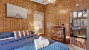 A bedroom with a wooden wall panel, a bed with blue bedding, a ceiling fan, a nightstand with a lamp, a dresser, and a window with blinds. - The Alpine Arena (Clifton Township)