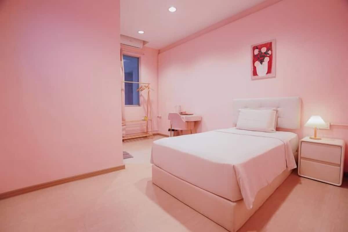 A pink-themed bedroom with a large bed, a bedside table with a lamp, and a small desk with a chair.
