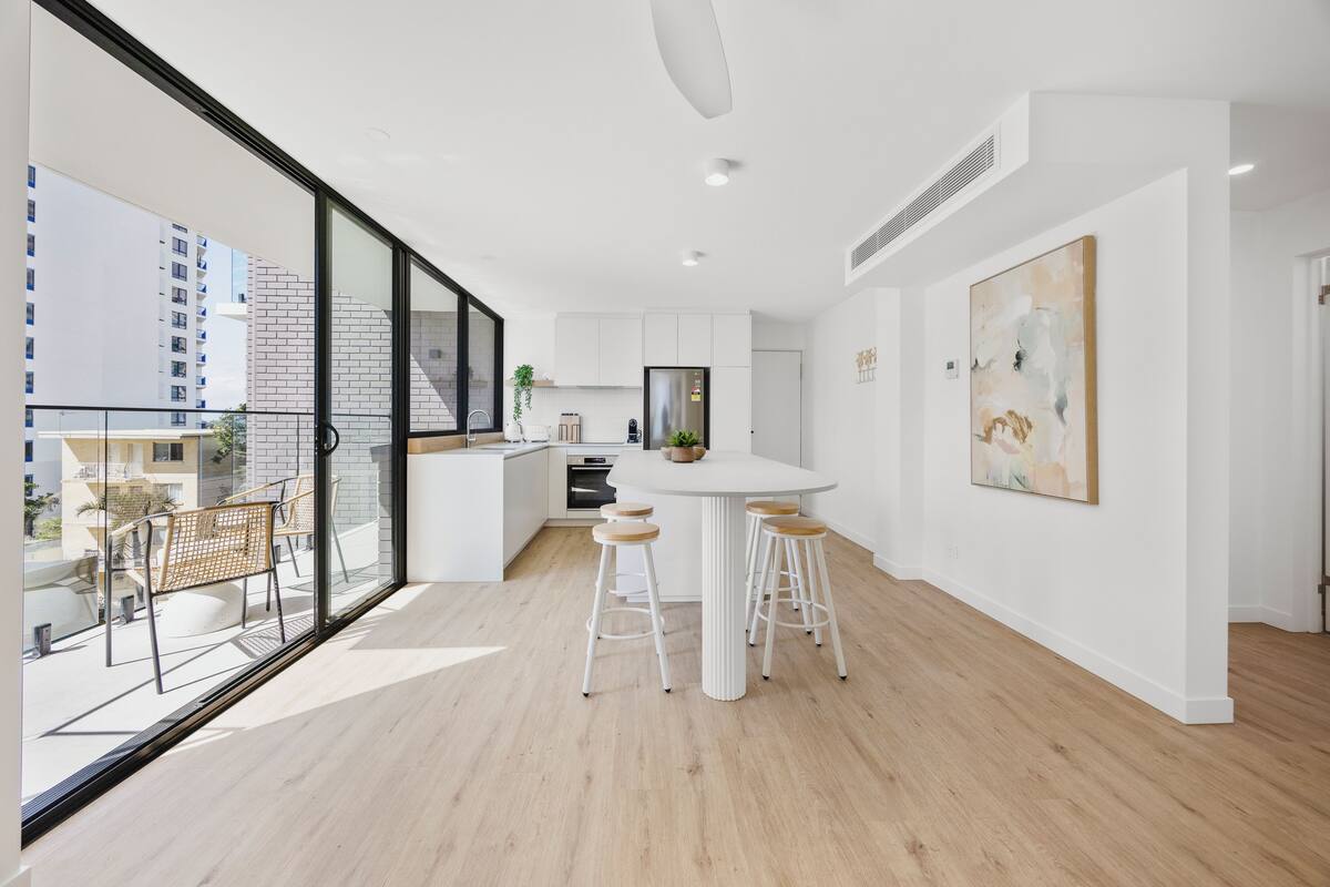 Una cocina moderna con armarios blancos, una isla central y taburetes. Hay un ventanal que ofrece vistas al exterior.
