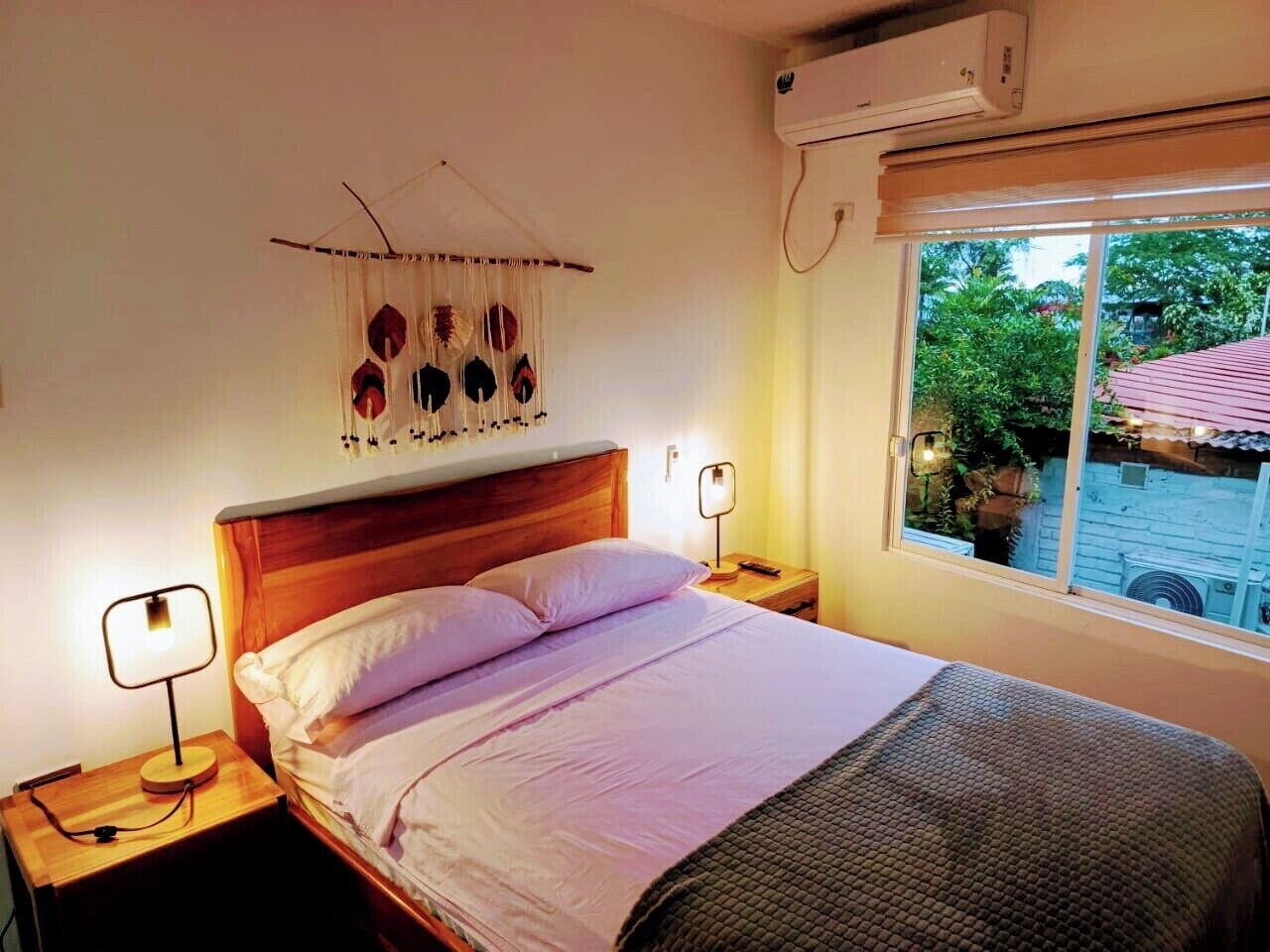 Een slaapkamer met een houten hoofdbord, een bed met witte lakens, twee nachtkastjes met lampen, een wandairconditioner, en een raam met uitzicht op groen.