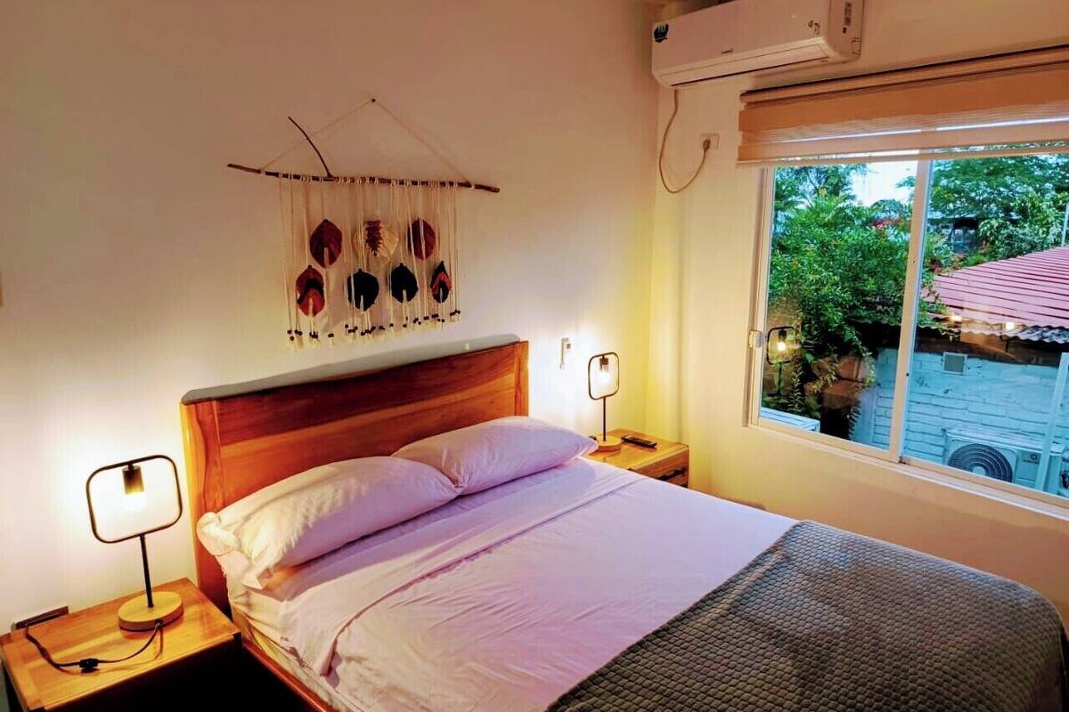 Un dormitorio con cabecera de madera, cama con ropa de cama blanca, dos mesitas de noche con lámparas, aire acondicionado de pared y ventana con vista a follaje.