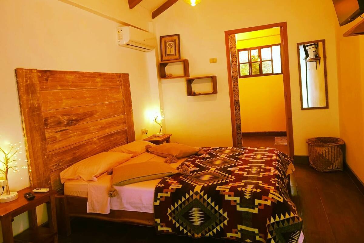 Un dormitorio con cabecera de madera, cama con manta estampada, lámpara de noche, aire acondicionado instalado en la pared y un espejo.
