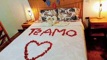 A bed with a floral bedspread, a heart-shaped arrangement of rose petals spelling "TE AMO", and a hanging decorative item.