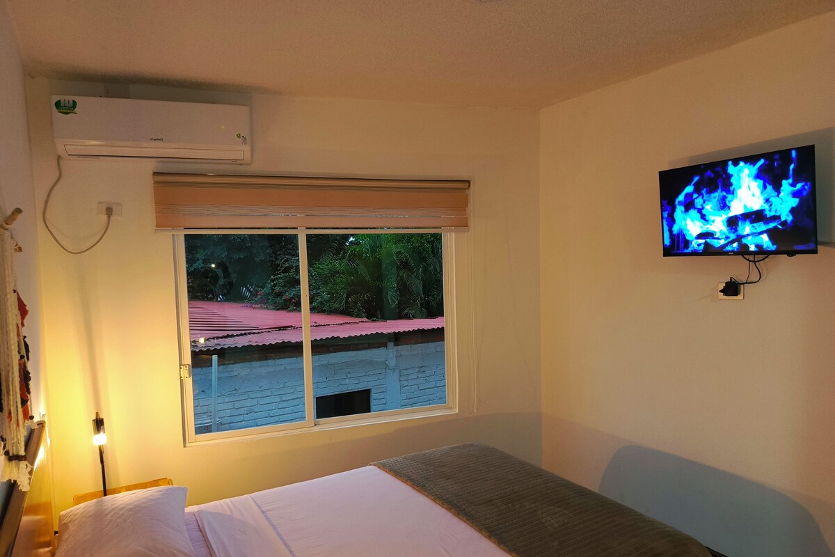 Una habitación de hotel con cama, ventana con vista a un edificio de techo rojo, televisor montado en la pared y unidad de aire acondicionado.