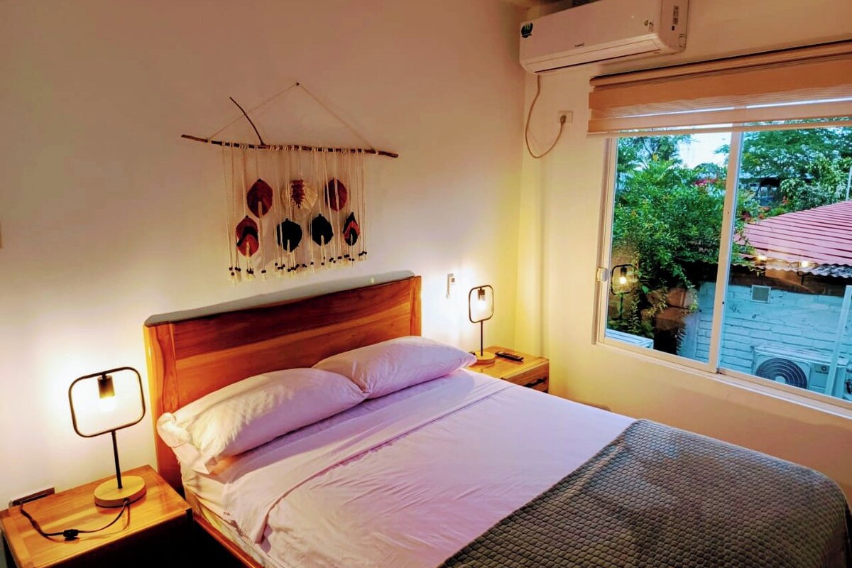 A bedroom with a wooden headboard, a bed with white linens, two bedside tables with lamps, a wall-mounted air conditioner, and a window with a view of greenery outside.