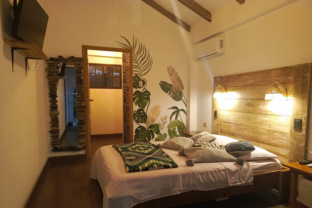 A bedroom with a wooden headboard, a bed with multiple pillows, a wall-mounted air conditioner, and a door leading to another room.