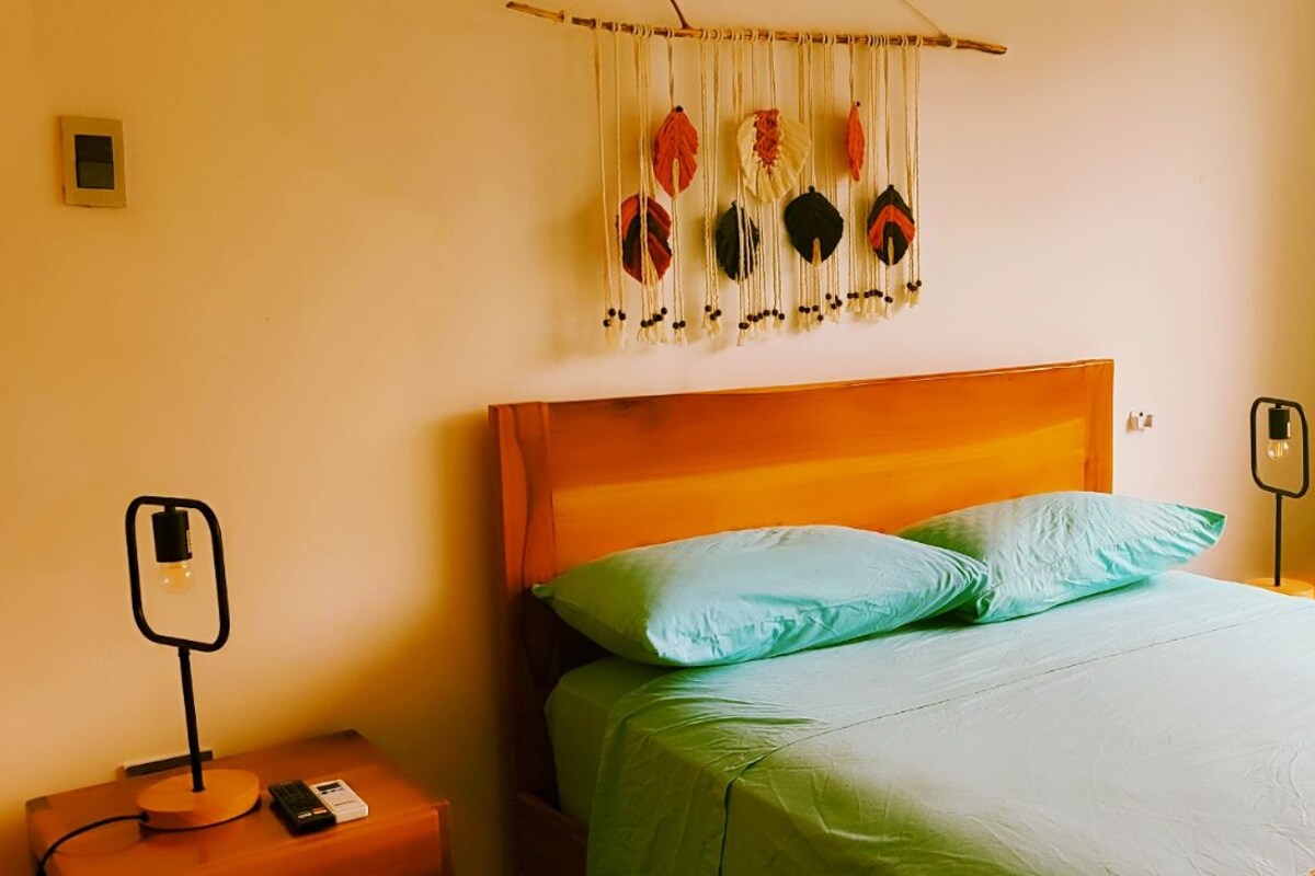 A bedroom with a bed, a bedside table, a wall-mounted light, a power outlet, and a window with a view of greenery outside.