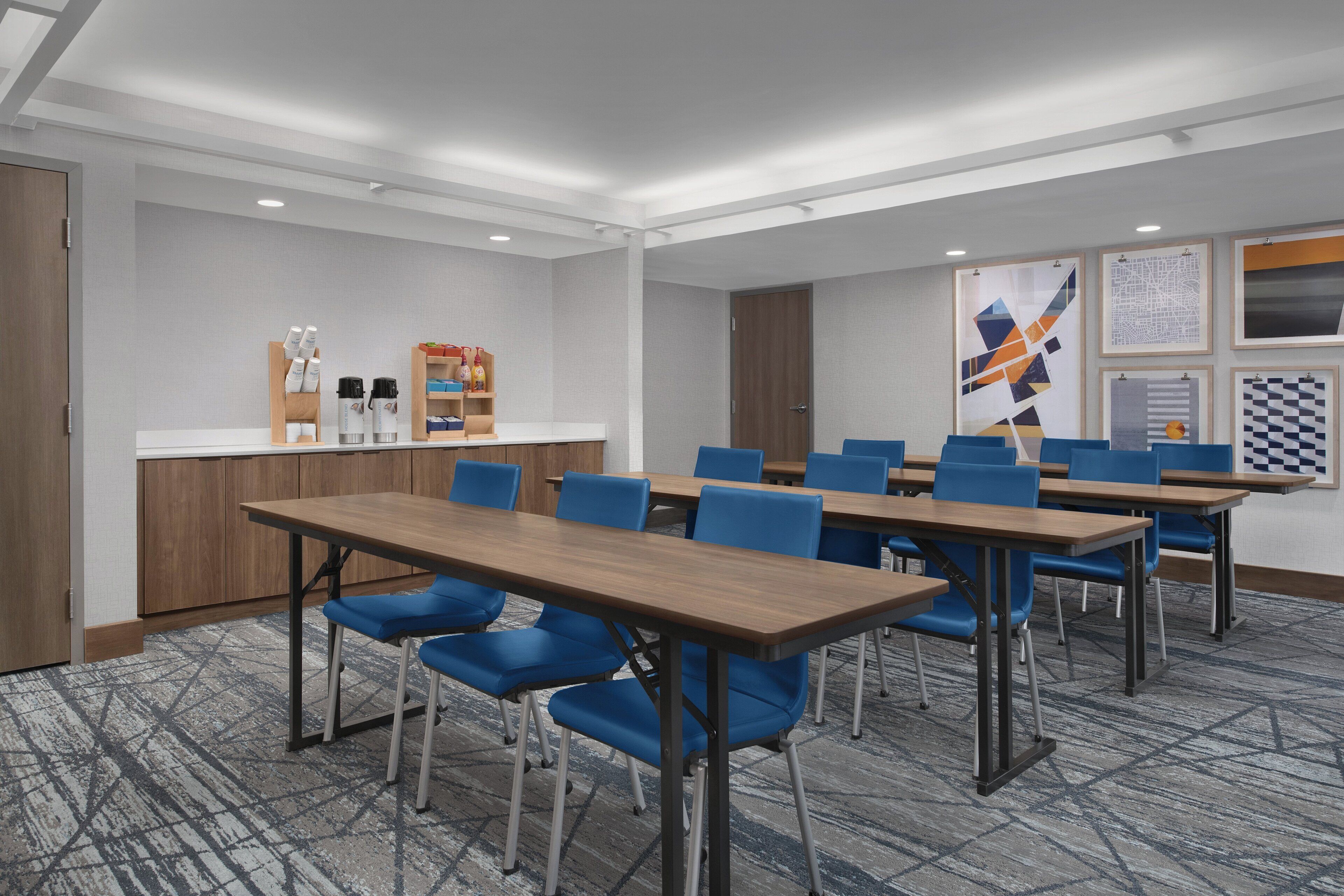 A conference room with multiple tables and blue chairs, a kitchenette area, and framed artwork on the walls.
