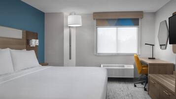 A hotel room with a large bed, a desk with a chair, a flat-screen TV, a standing lamp, and a window with blinds.