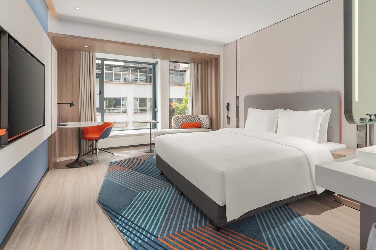 Une chambre d’hôtel moderne avec un grand lit, un bureau et une chaise. Elle est équipée d’une fenêtre avec des rideaux, d’une salle de bain avec un miroir et d’un téléviseur fixé au mur.