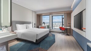 A modern hotel room with a large bed, a desk, a chair, a TV, and a view of the city. - Holiday Inn Express Puer Simao by IHG (Pu'er)