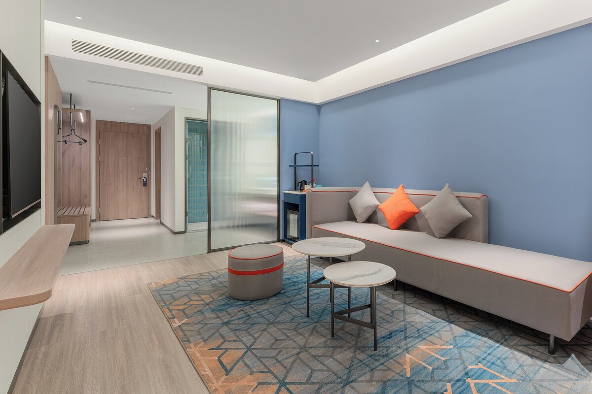 A modern hotel room with a sofa, a small table, a bench, and a desk with a lamp.