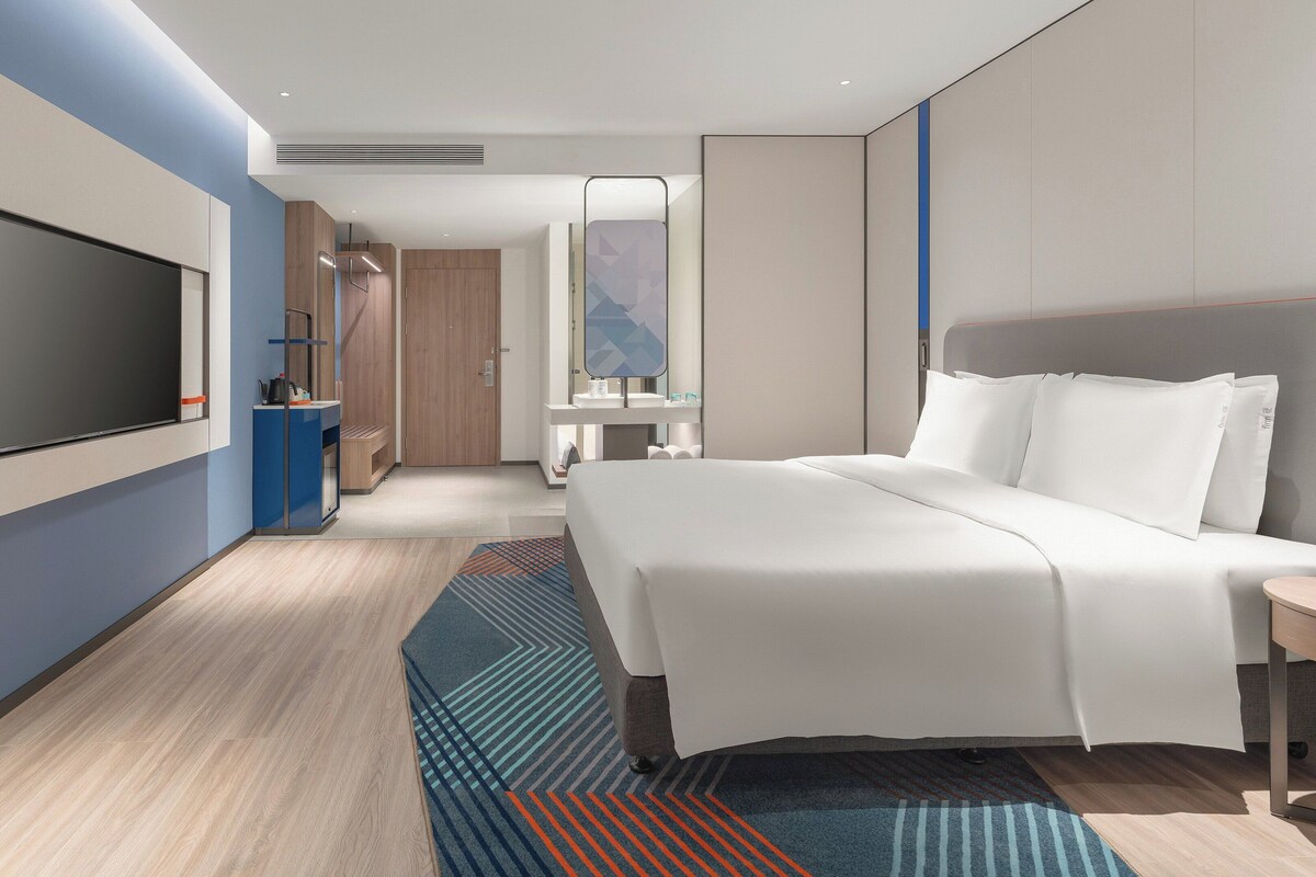 A modern hotel room with a large bed, a flat-screen TV, a bedside table, and a bathroom with a mirror.