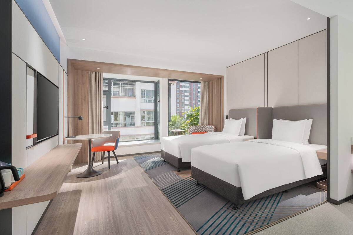 Une chambre d’hôtel moderne équipée de deux lits, d’un bureau, d’une chaise, d’une grande fenêtre et d’une salle de bain avec un miroir et un porte-serviettes.