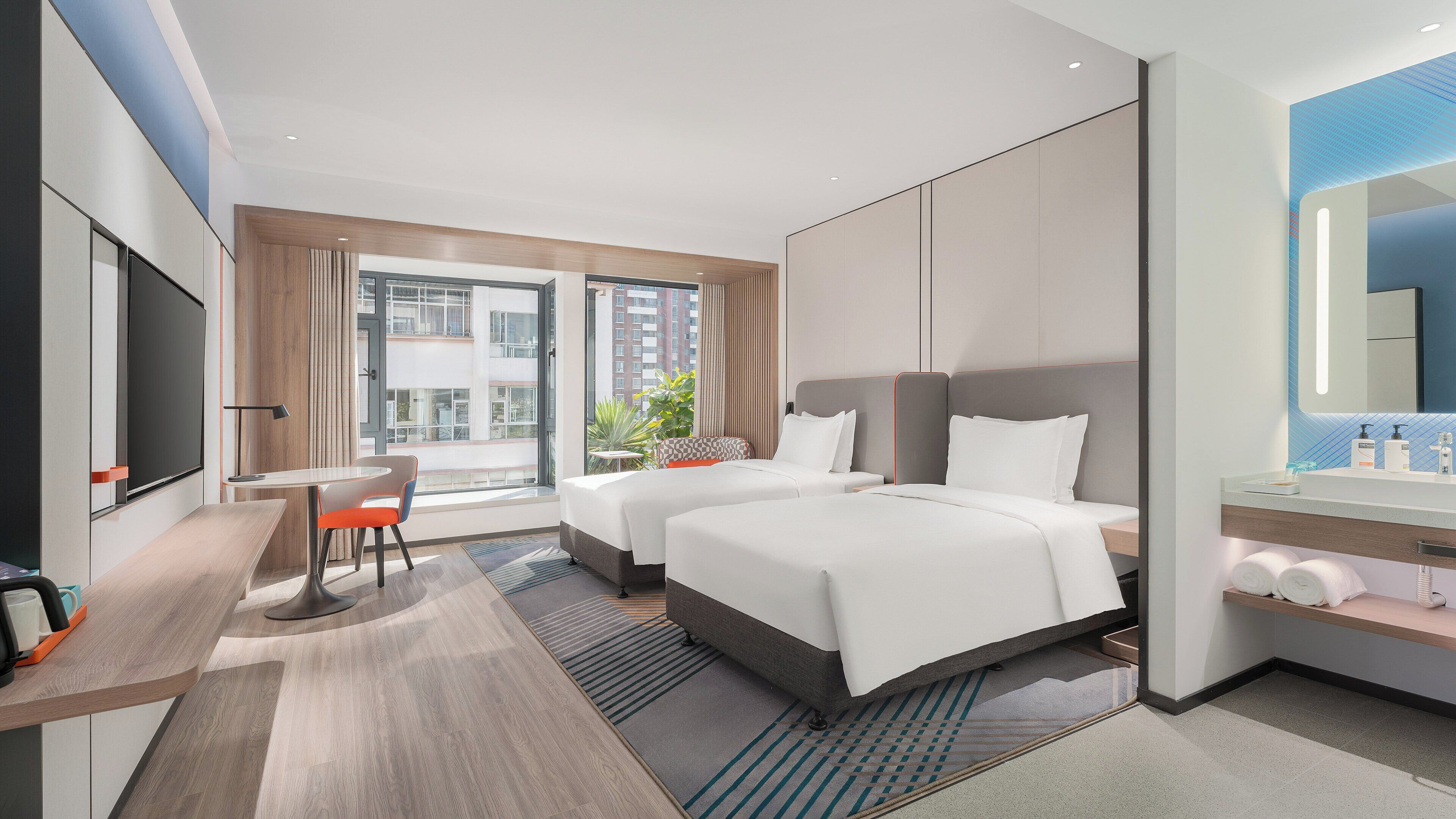 Habitación de hotel moderna con dos camas, un escritorio, una silla, un ventanal grande y un baño con espejo y toallero.