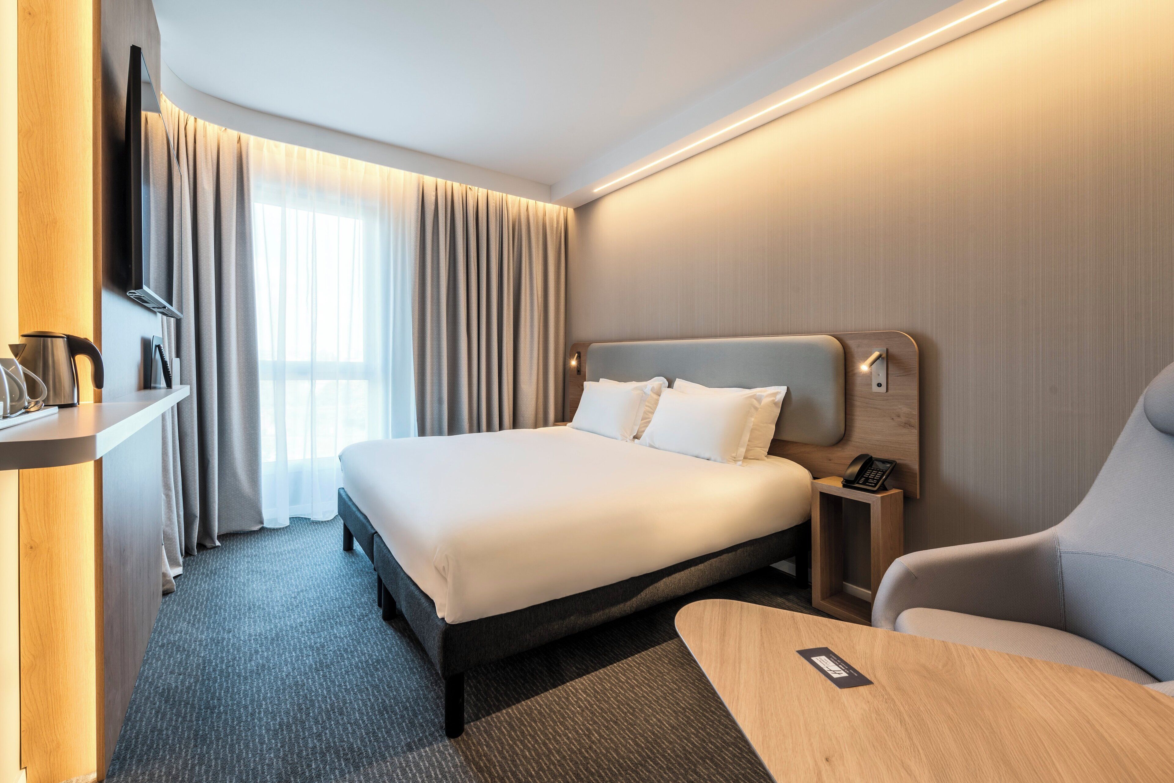 A hotel room with a large bed, a desk with a phone, a chair, a TV, and a window with curtains.