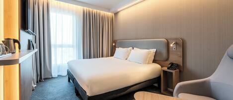 A hotel room with a large bed, a desk with a phone, a chair, a TV, and a window with curtains.