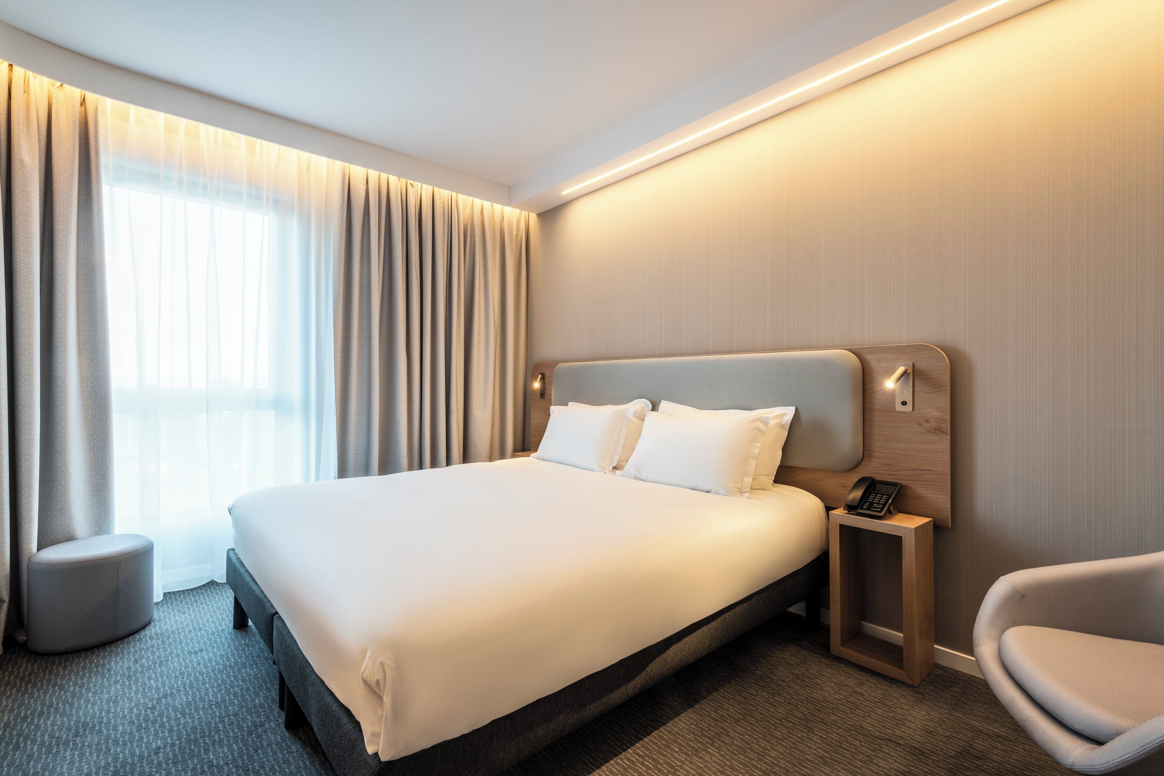 A hotel room with a large bed, a bedside table with a phone, a chair, and a window with curtains.