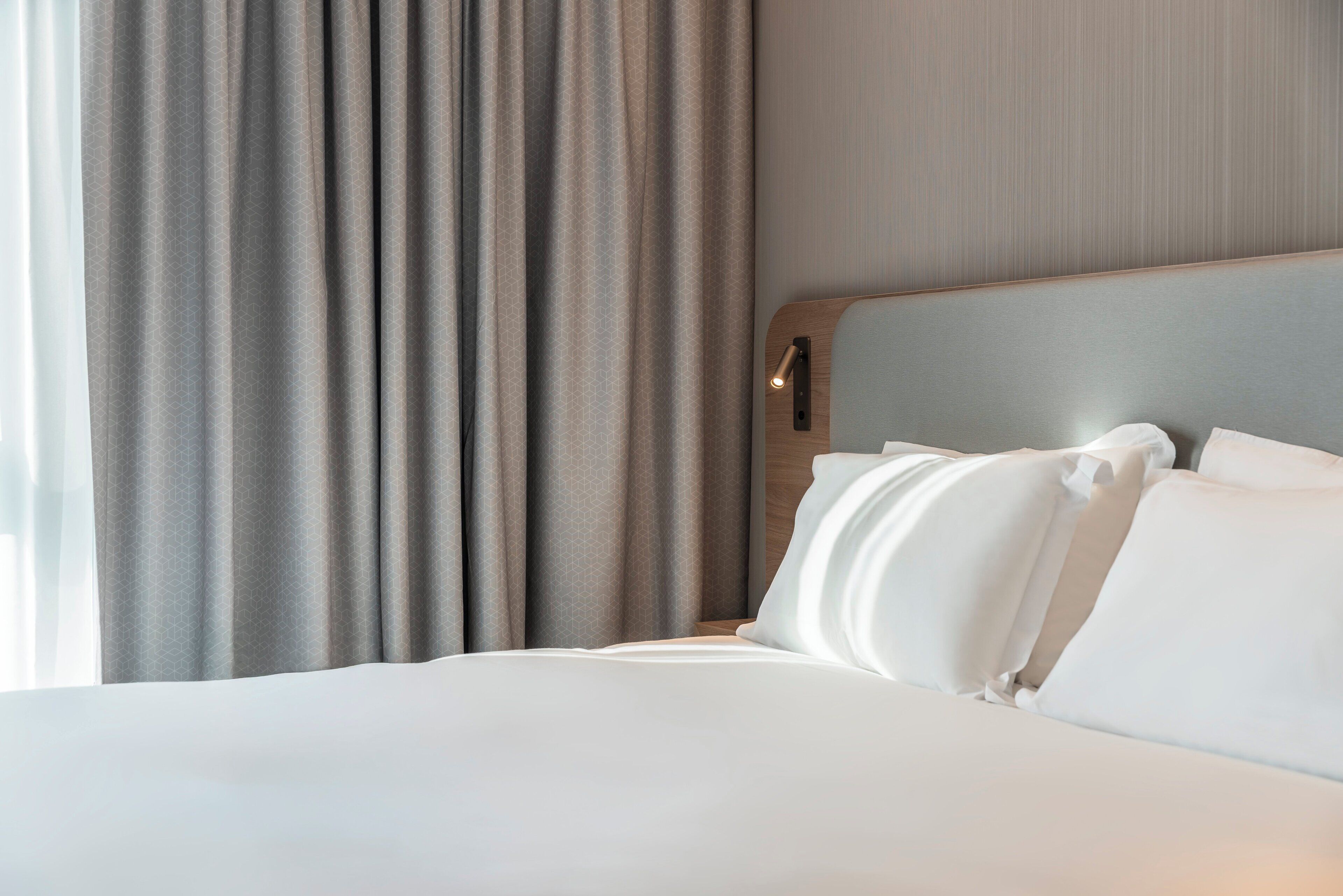 A hotel room with a neatly made bed, a wooden headboard, and a large window with curtains.