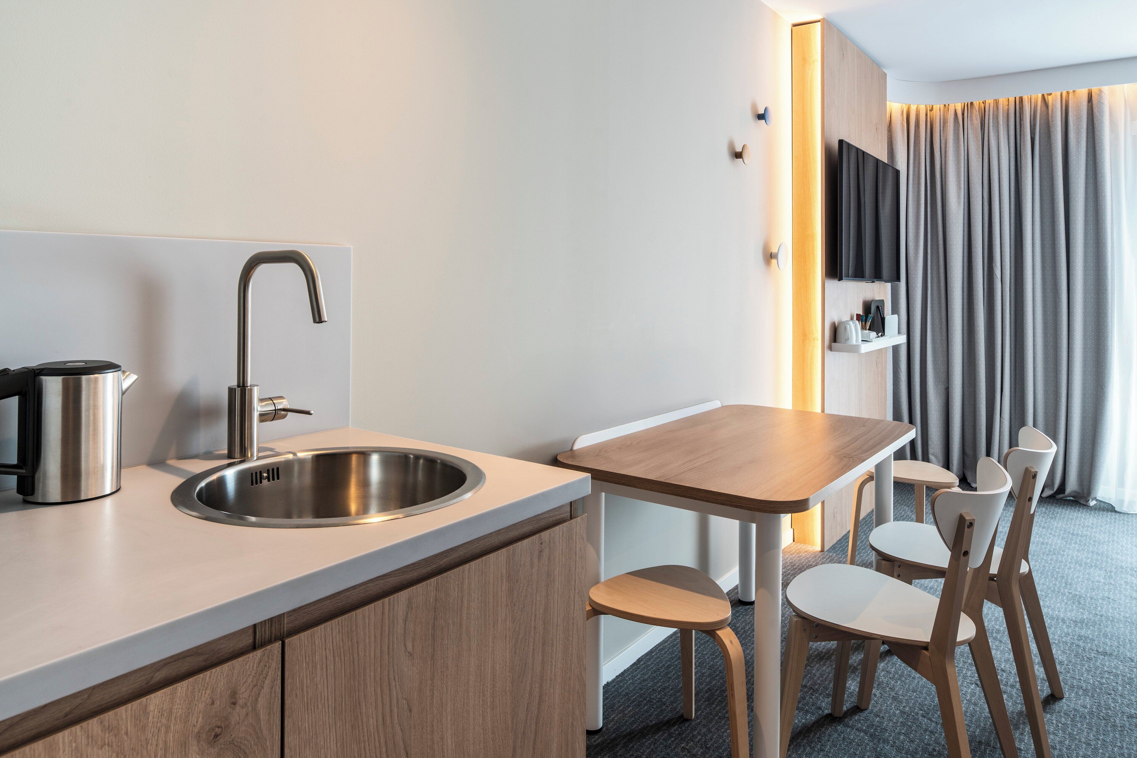 A modern hotel room with a kitchenette, a sink, a wooden table, and a TV mounted on the wall.