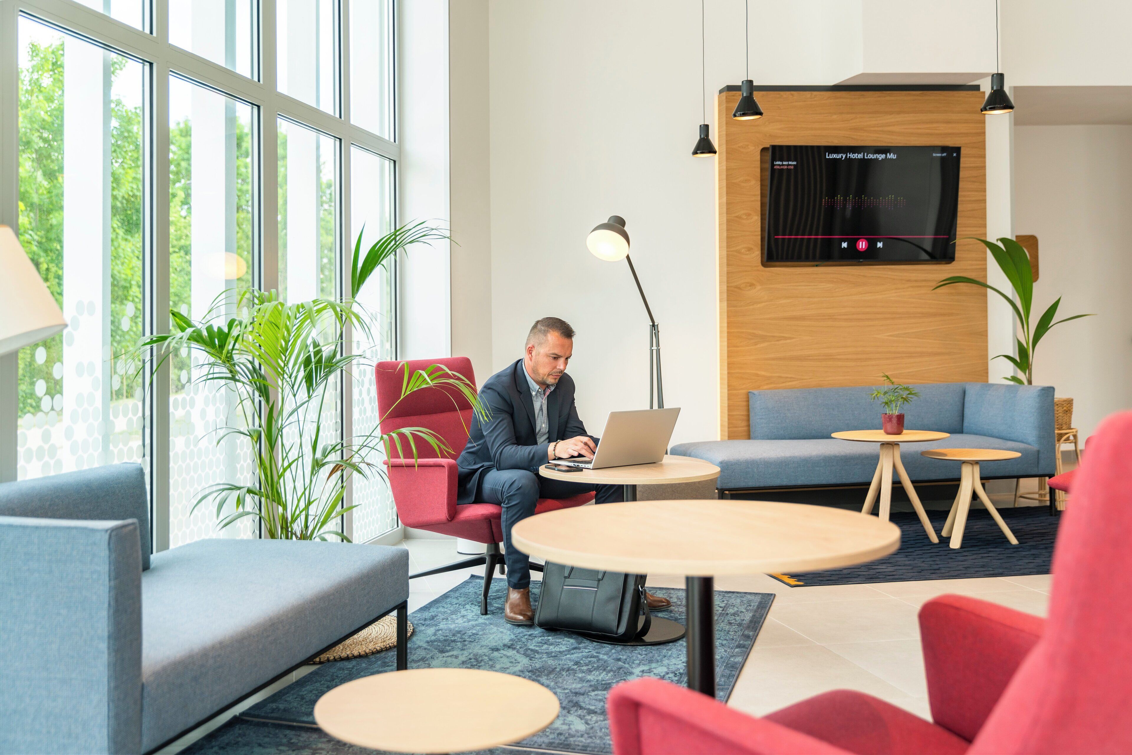 Un salón moderno de hotel con un hombre trabajando en un ordenador portátil, ventanas amplias y un televisor montado en la pared.