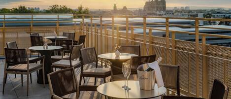 A terrace with tables and chairs, a champagne bucket, and a view of a city skyline with a prominent cathedral.