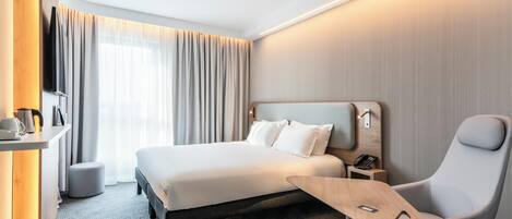 A modern hotel room with a large bed, a desk with a chair, a small table, and a telephone.