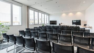 Una sala de conferencias con filas de sillas negras, una pantalla grande y una planta.