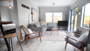 A living room with a sofa, two armchairs, a coffee table, a TV, and a large window with blinds. - Quiet Escape with Lake Access and Kayaks | Peaceful Cosy Cottage (Rotorua)