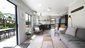 A modern living room with a grey sofa, a coffee table, and a dining area with a table and chairs. There is a kitchen area with a fridge and a sink. - Quiet Escape with Lake Access and Kayaks | Peaceful Cosy Cottage (Rotorua)