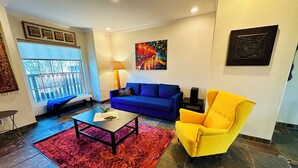 A living room with a blue sofa, a yellow armchair, a coffee table, a red rug, and a large window with a view.