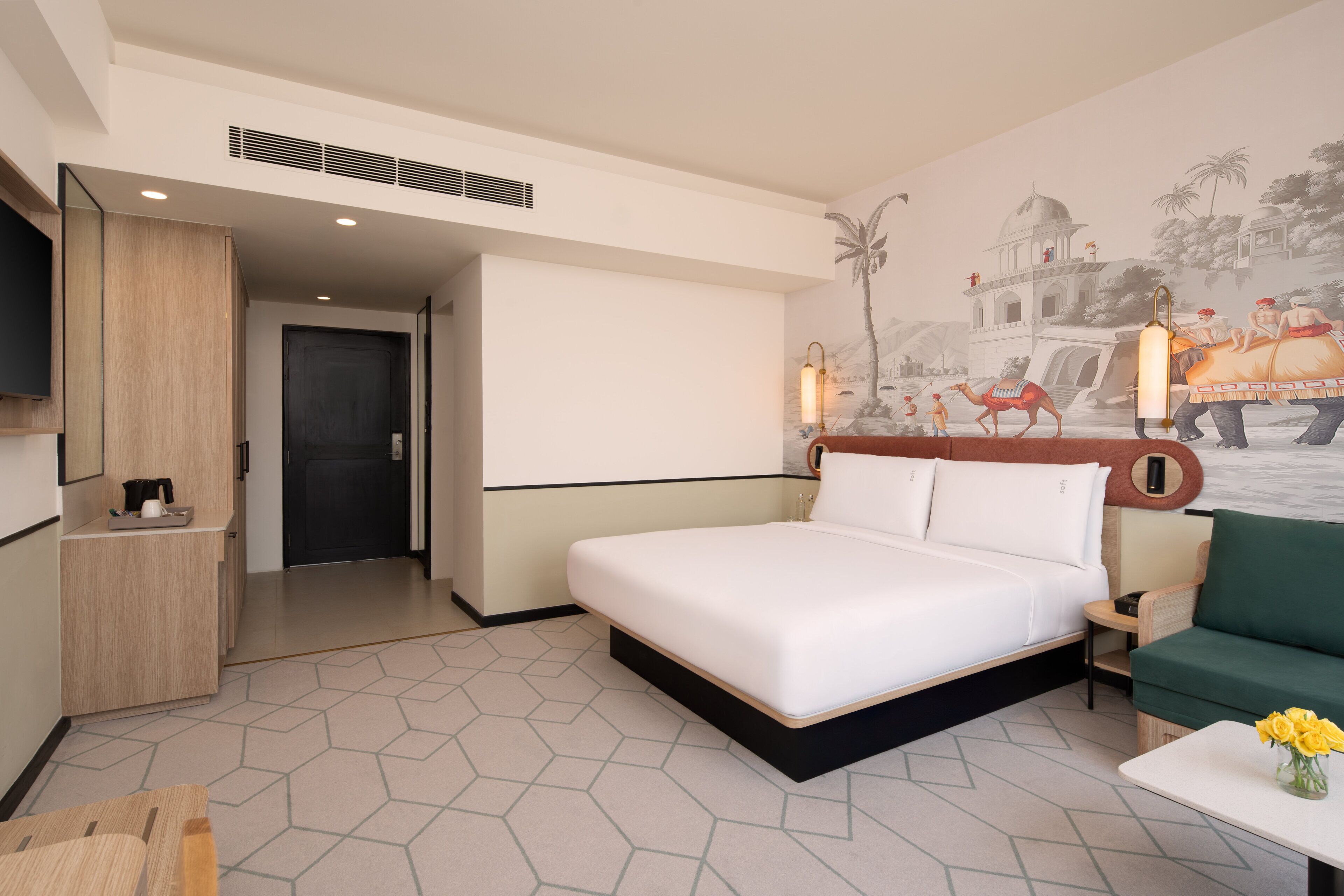 A hotel room with a bed, a green armchair, a small table with flowers, a TV, and a wall mural.