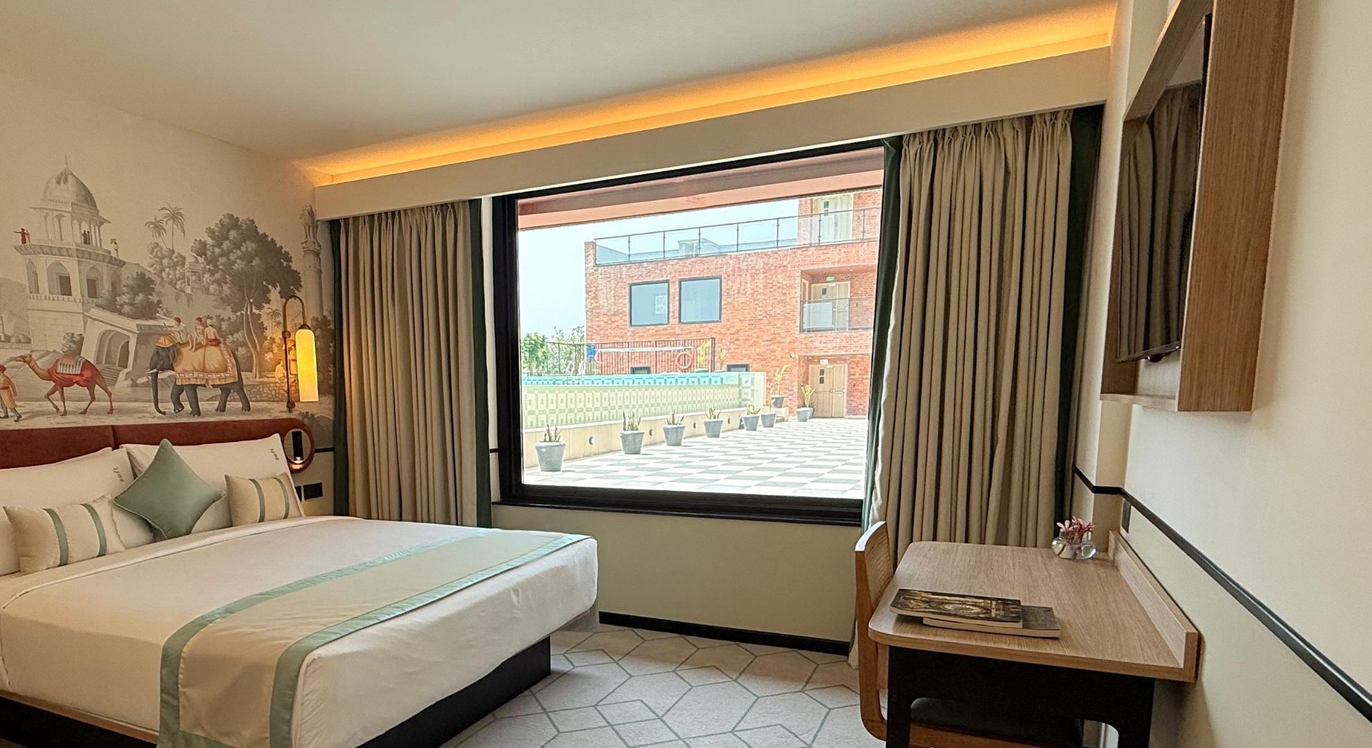 A hotel room with a large bed, a desk, a window with curtains, and a mural on the wall.